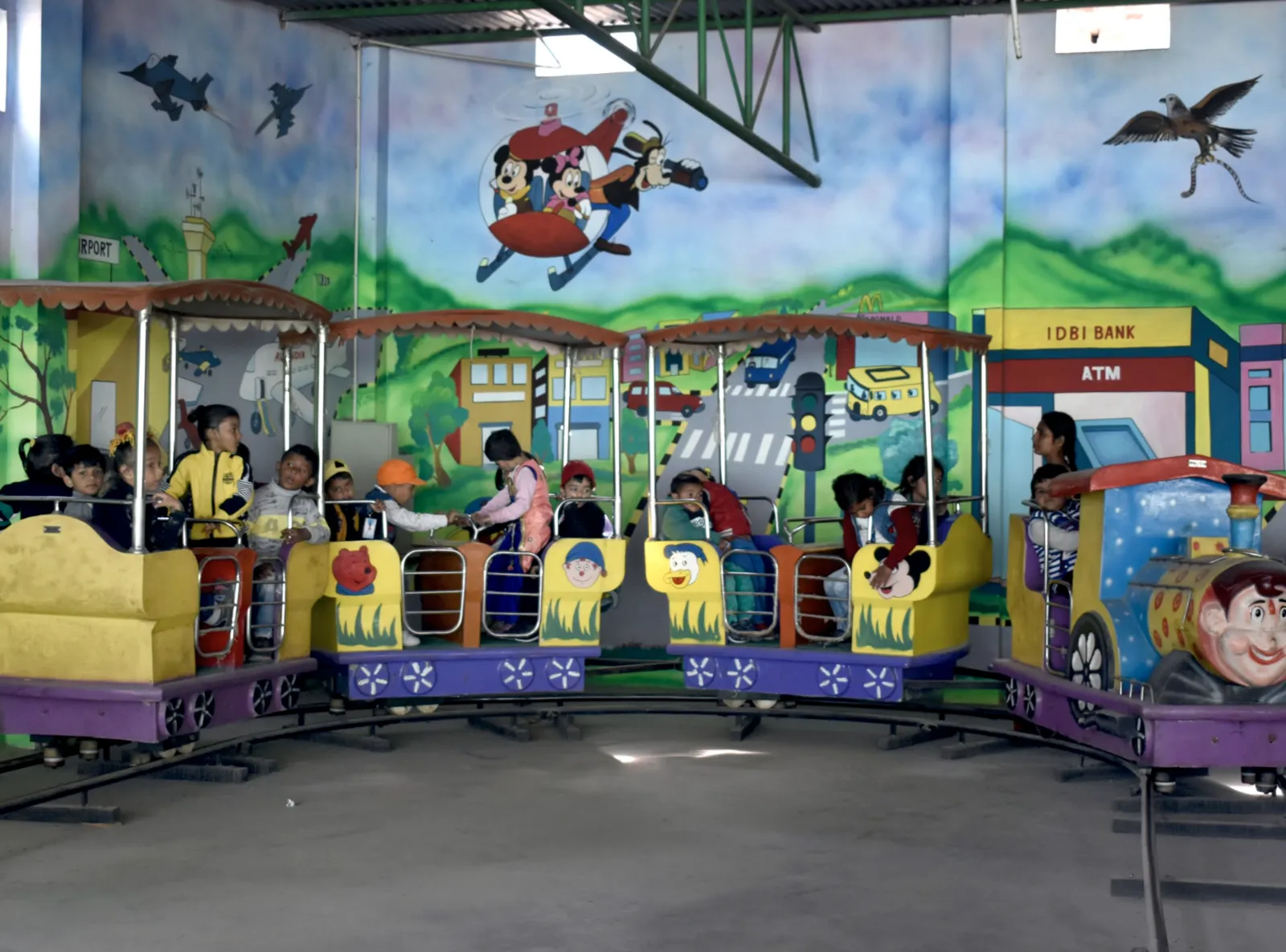 Young children riding the toy train at the KG Wing, in front of a painted cartoon mural