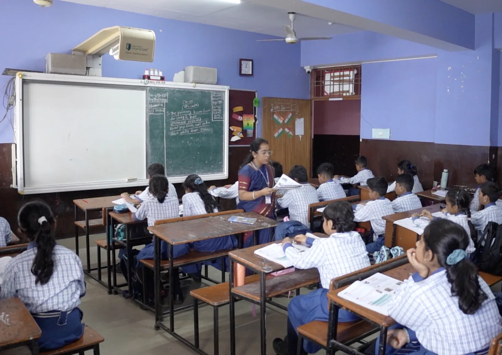 A teacher handing out worksheets in a smart classroom with a digital board beside the blackboard