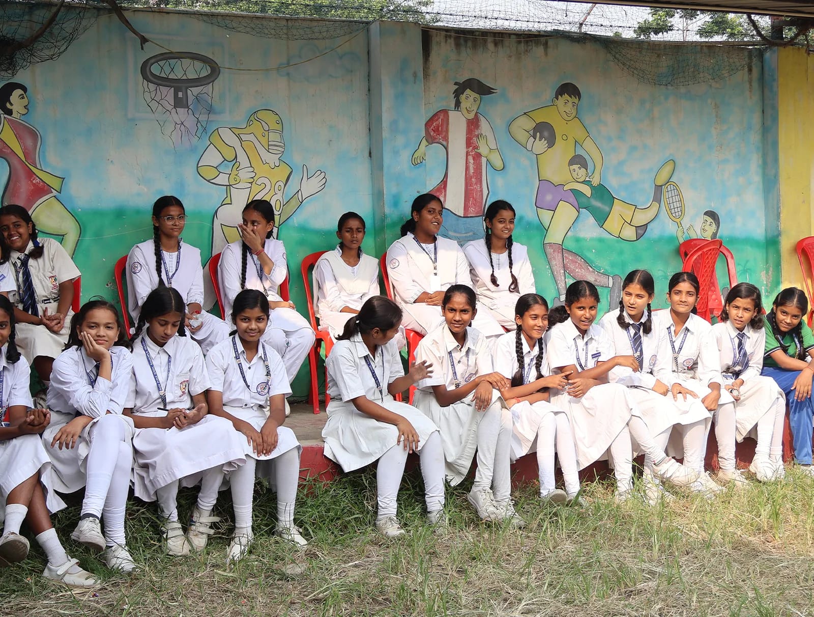 Girls in white uniforms sitting in front of the sports mural, cheering the cricket matches