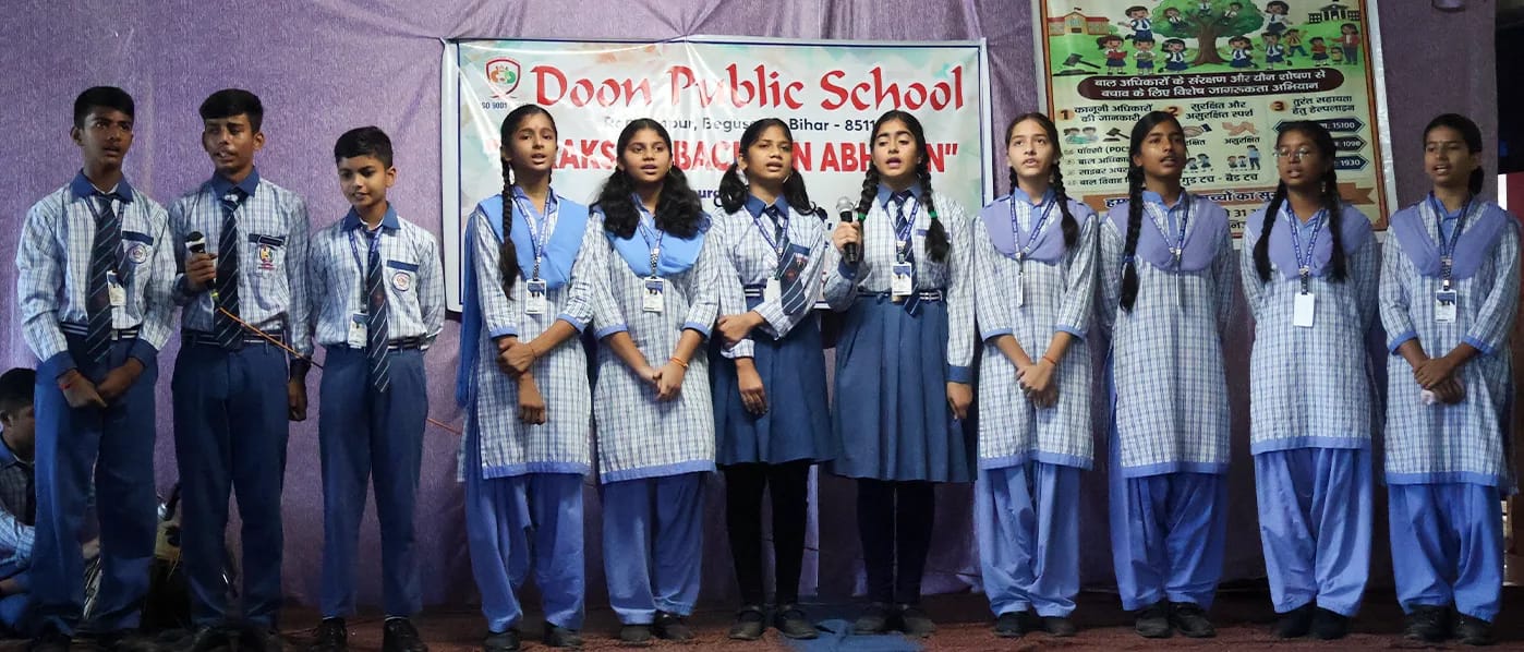 Students standing in a line on stage in front of the Surakshit Bachpan Abhiyan banner